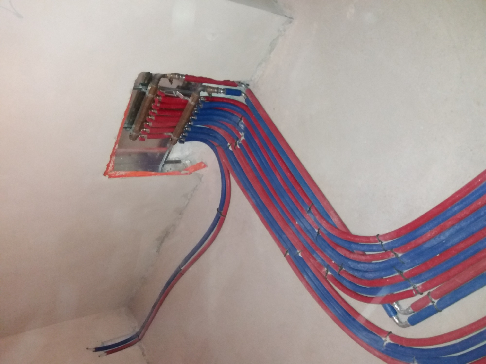 Rozprowadzenie instalacji ogrzewania podłogowego: czerwone i niebieskie rury prowadzone od rozdzielacza w ścianie, widoczne mocowania i surowy stan wykończenia.