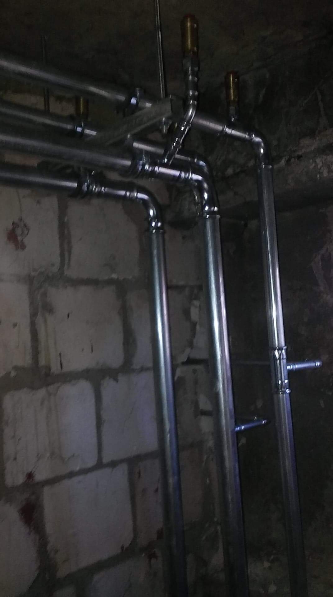 Metalowe rury instalacji hydraulicznej, prowadzone wzdłuż surowej ściany z cegieł, z widocznymi zaworami i mocowaniami.