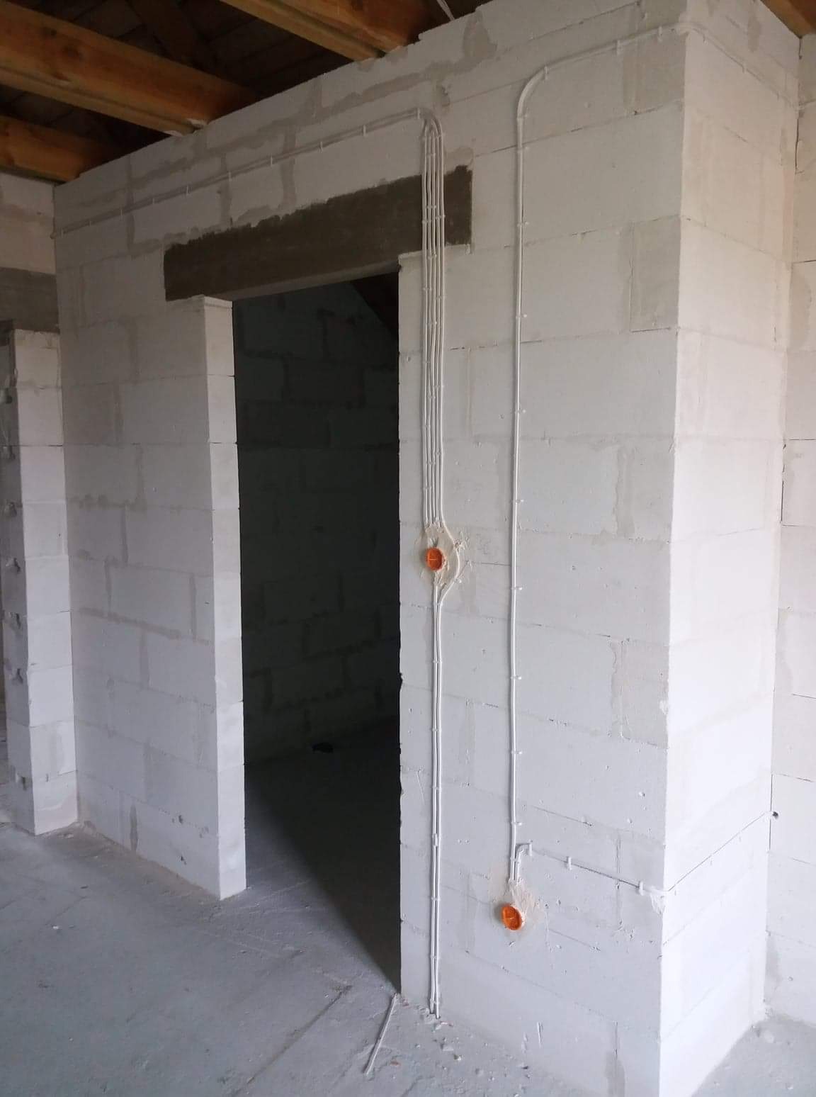 Wewnętrzna instalacja elektryczna w surowym budynku z białych bloczków, widoczne kable w peszlach i pomarańczowe puszki podtynkowe.