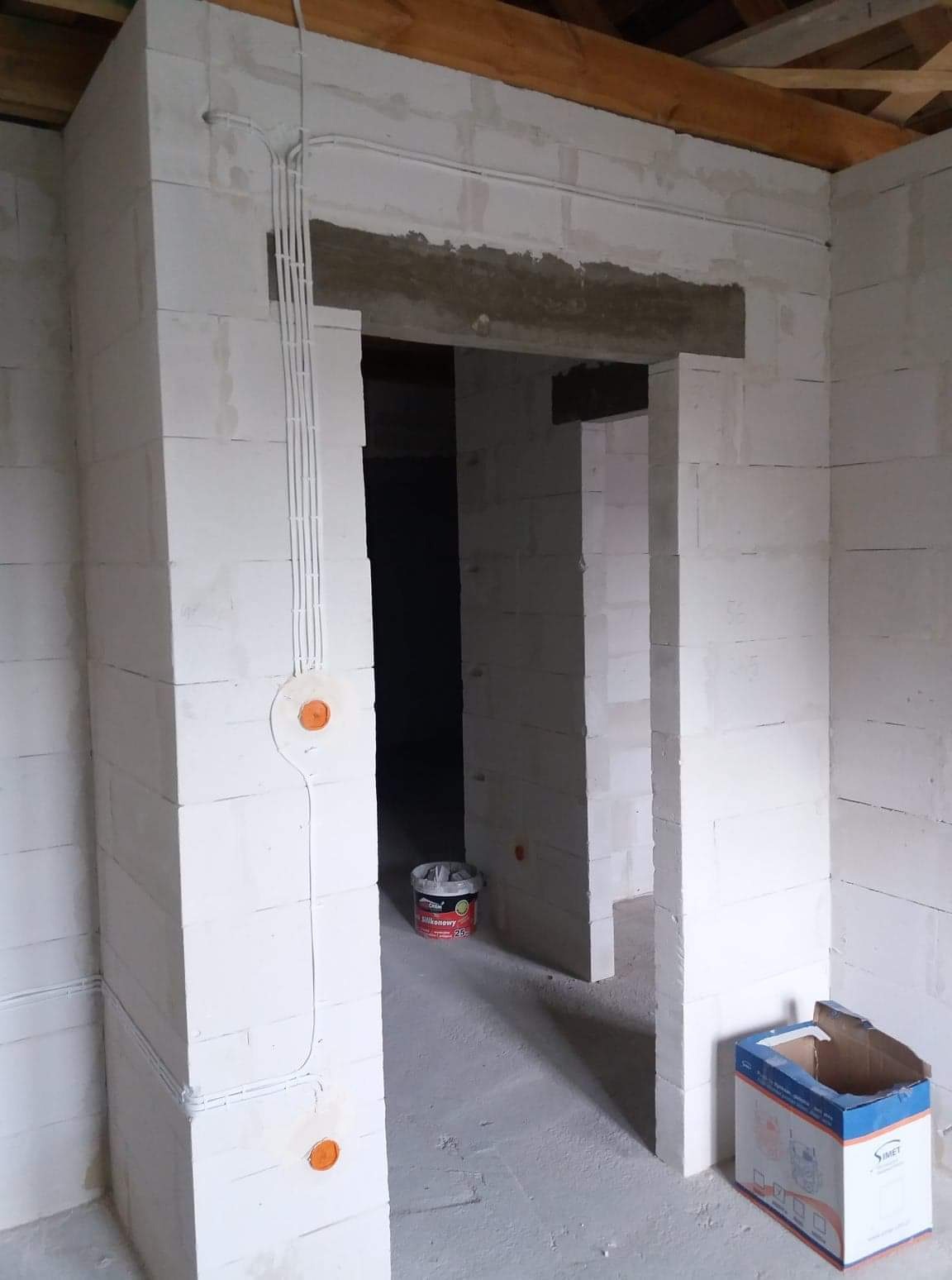 Wnętrze w trakcie budowy: ściany z białych bloczków, widoczne kable elektryczne w peszlach na ścianie, puszki instalacyjne, nadproże z betonu, puste wiadro po farbie i kartonowe pudełko na podłodze.