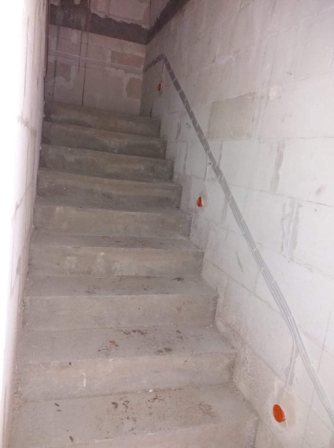 Surowe betonowe schody w trakcie budowy z widocznymi przewodami elektrycznymi w peszlu przy ścianie z bloczków betonowych.