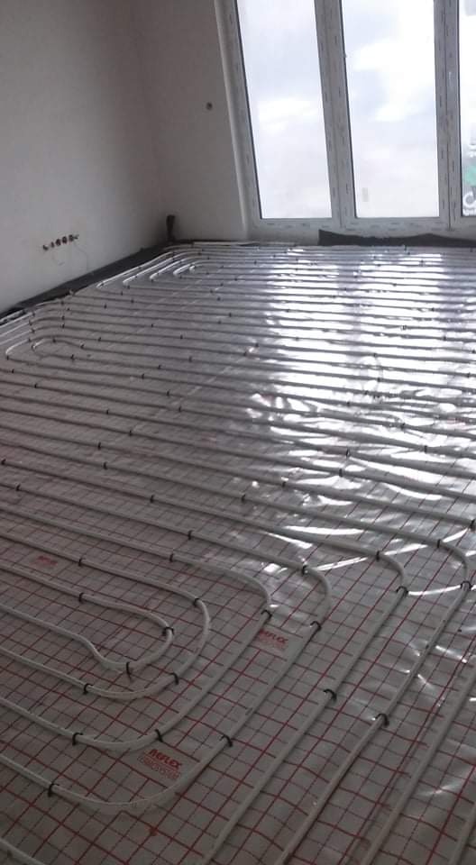 Białe rury ogrzewania podłogowego ułożone na macie grzewczej z siatką w odcieniach czerwieni, widoczne okno dachowe w jasnym pomieszczeniu.