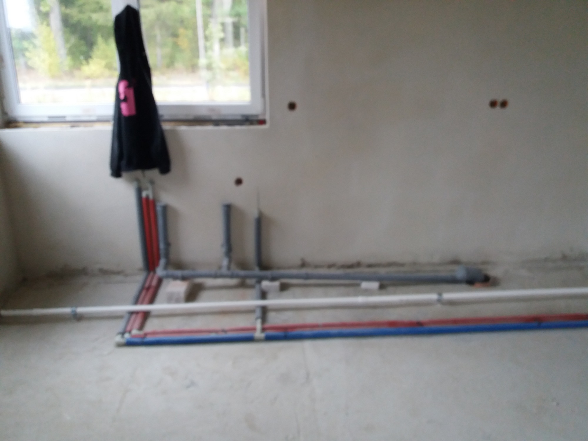 Surowe wnętrze z instalacją hydrauliczną w trakcie montażu: widoczne rury z tworzyw sztucznych w kolorach szarym, białym, czerwonym i niebieskim ułożone na podłodze, z fragmentami wystającymi ze...