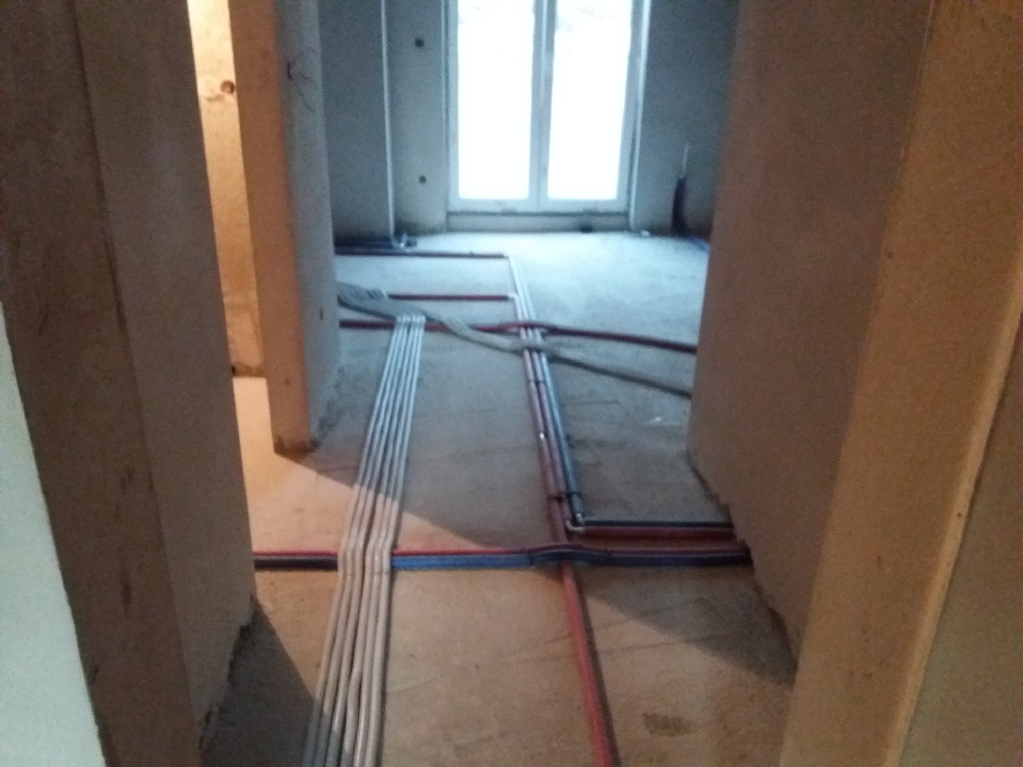 Wnętrze w trakcie remontu z widocznymi rurami instalacji wodnej i grzewczej ułożonymi na podłodze, przygotowanymi do zalania wylewką.