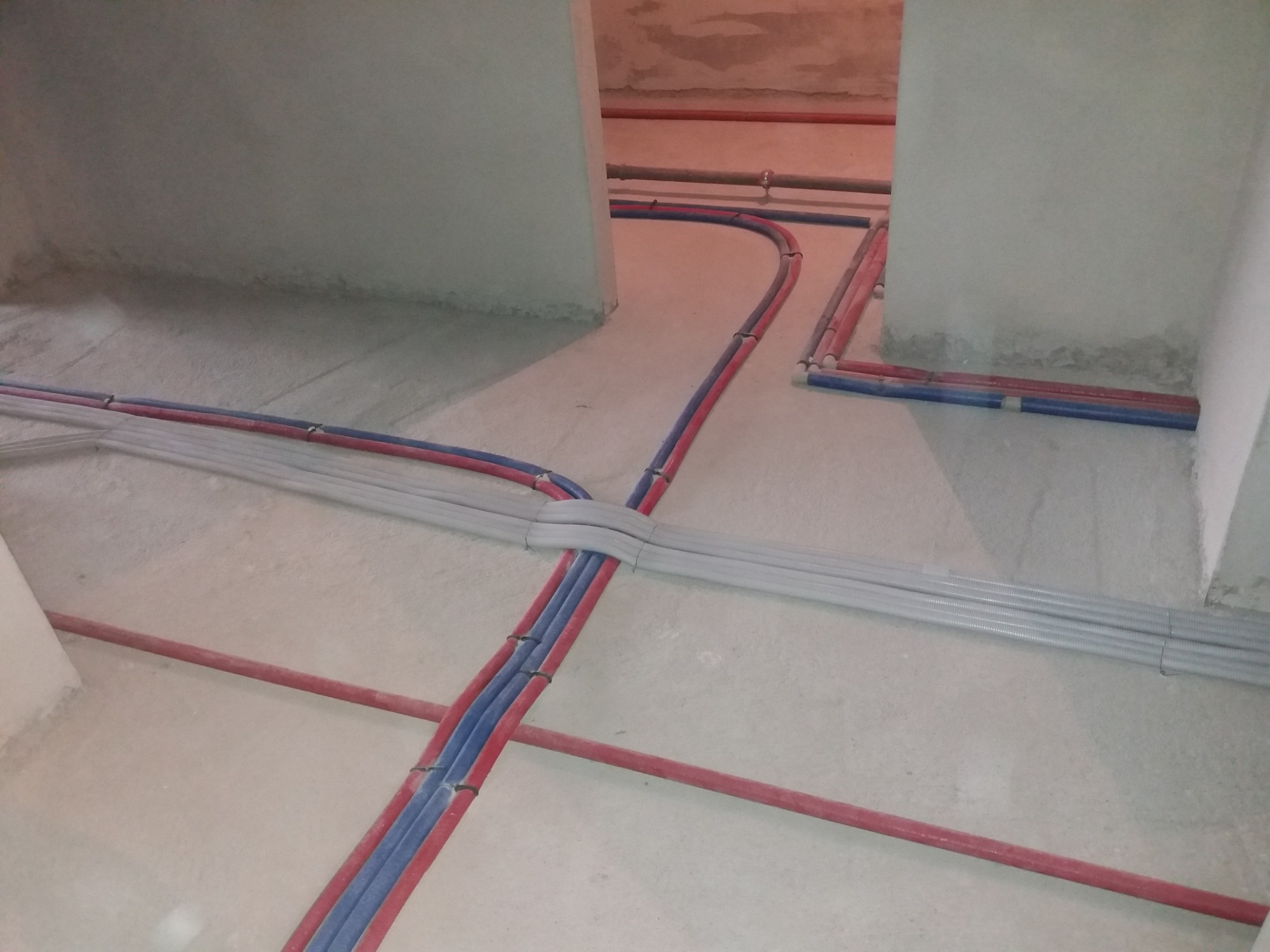 Rozprowadzone rury instalacji wodnej (czerwone i niebieskie) i szare rury karbowane na surowym podłożu wewnątrz budynku w trakcie budowy lub remontu.