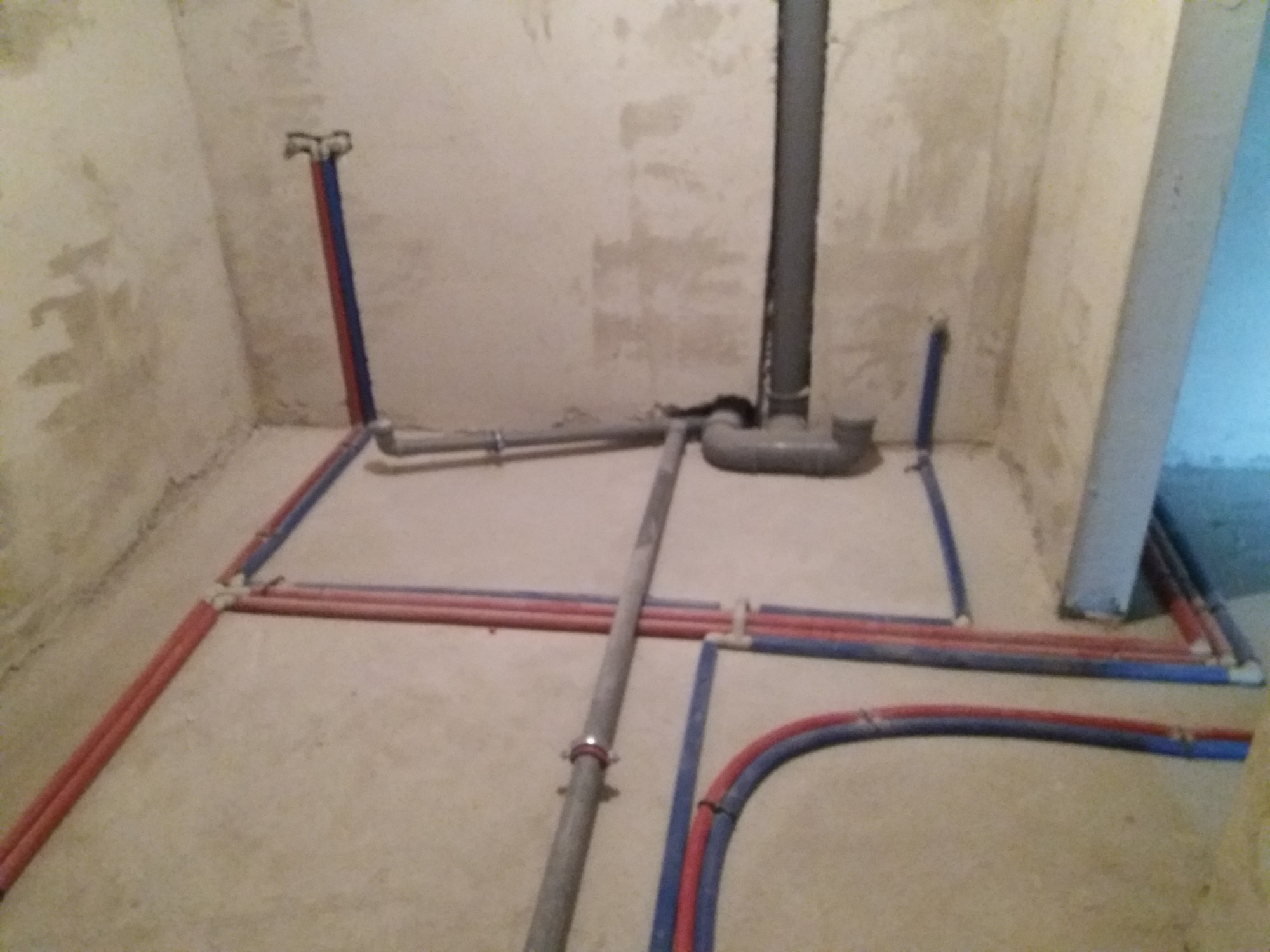 Surowy stan łazienki z rozprowadzonymi rurami wodnymi (czerwone i niebieskie) oraz szarą rurą kanalizacyjną pionową i poziomą, przygotowanymi do dalszych prac wykończeniowych.