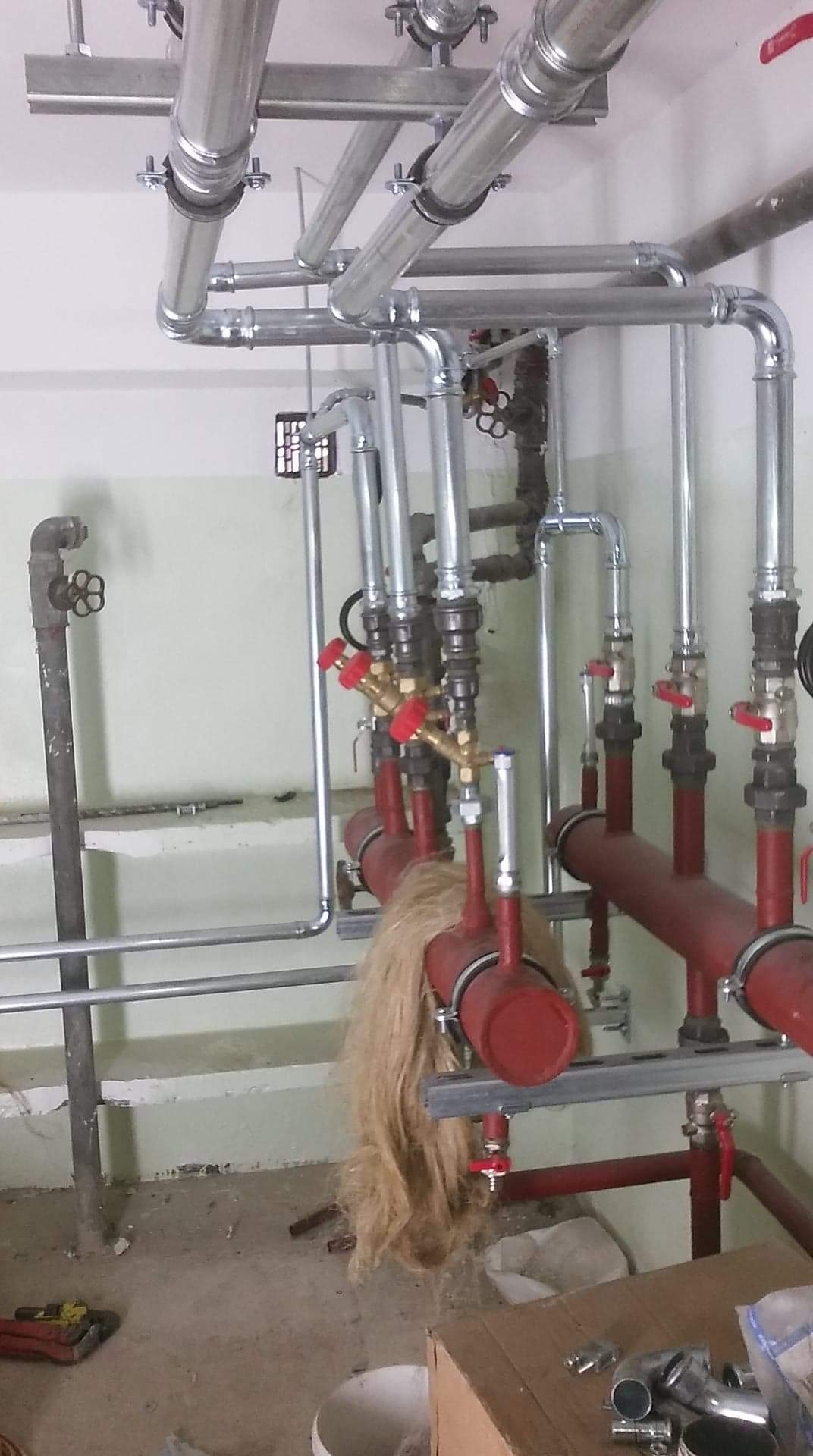Rozbudowana instalacja grzewcza z rur stalowych i miedzianych zaworów, częściowo owinięta izolacją z naturalnego włókna, zamontowana w pomieszczeniu technicznym z surowym betonowym podłożem.