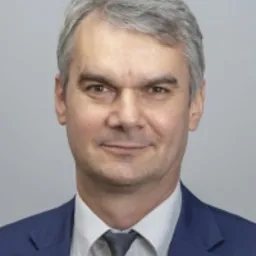 Portret biznesowy mężczyzny w średnim wieku, z siwiejącymi włosami, w niebieskim garniturze i szarym krawacie, na jasnoszarym tle. Uśmiech nieznaczny, wzrok skierowany wprost.