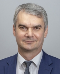 Portret biznesowy mężczyzny w średnim wieku, z siwiejącymi włosami, w niebieskim garniturze i szarym krawacie, na jasnoszarym tle. Uśmiech nieznaczny, wzrok skierowany wprost.