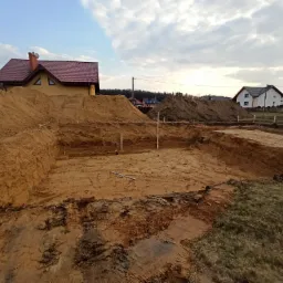 Świeżo wykopany fundament pod budynek, z widocznymi stertami ziemi, drewnianymi słupkami wyznaczającymi obrys i domami w tle, sfotografowany w pochmurny dzień.