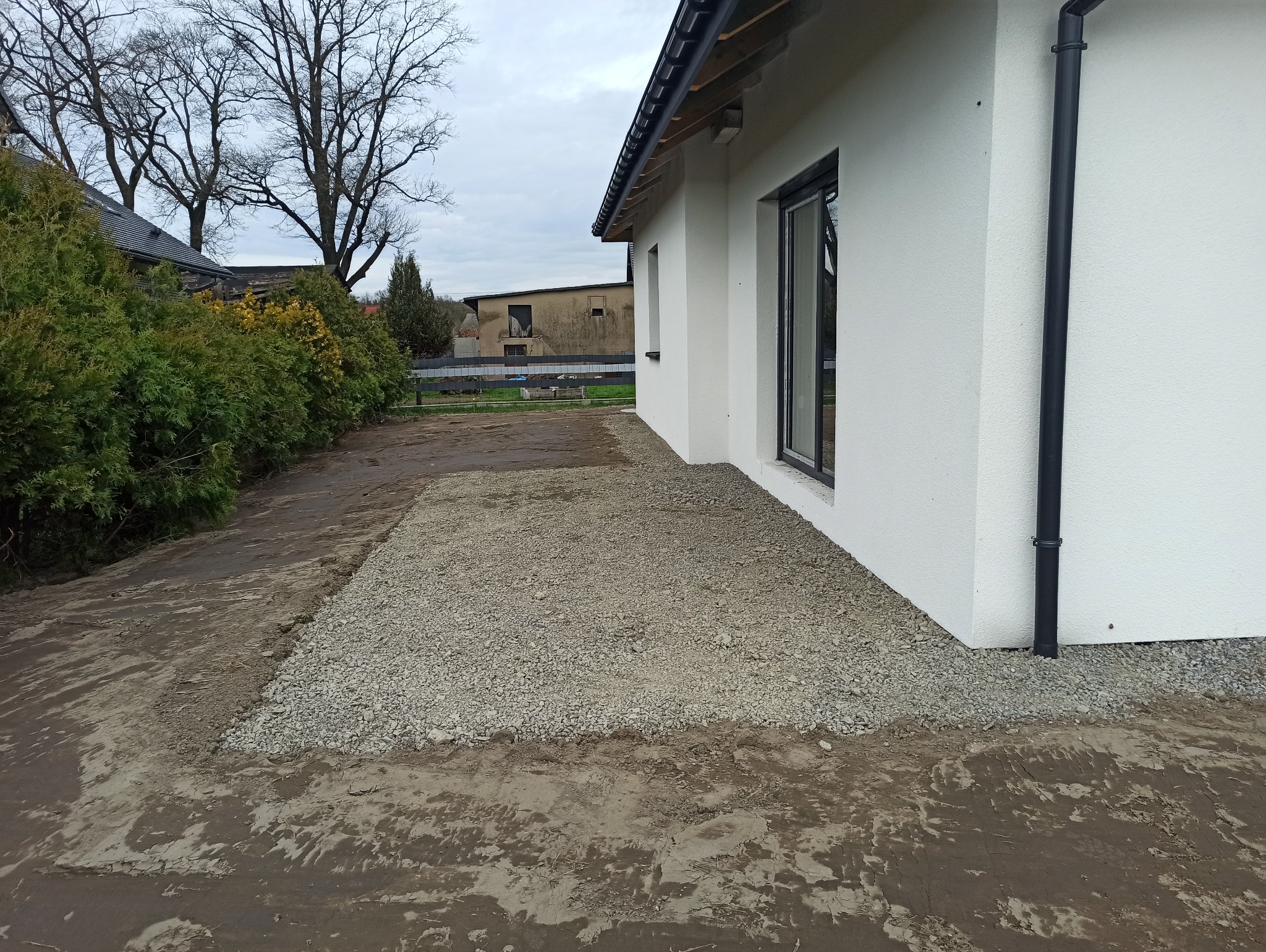 Przygotowanie terenu pod taras: utwardzona, kamienista powierzchnia przylegająca do białej ściany domu z czarną rynną, widoczne przygotowane podłoże z humusem, w tle zieleń i zabudowania.