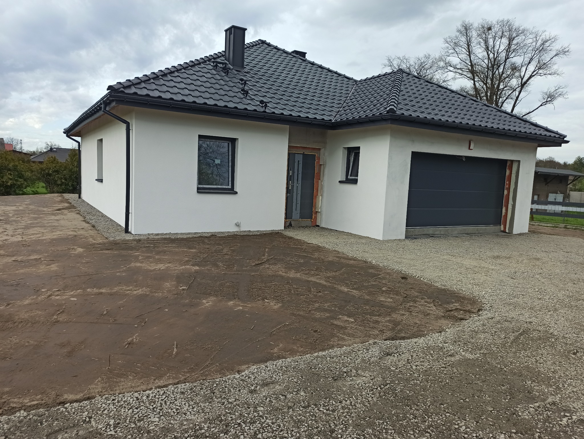 Nowoczesny dom jednorodzinny z grafitową dachówką i jasną elewacją, teren wokół domu częściowo utwardzony żwirem, a częściowo przygotowany pod trawnik.