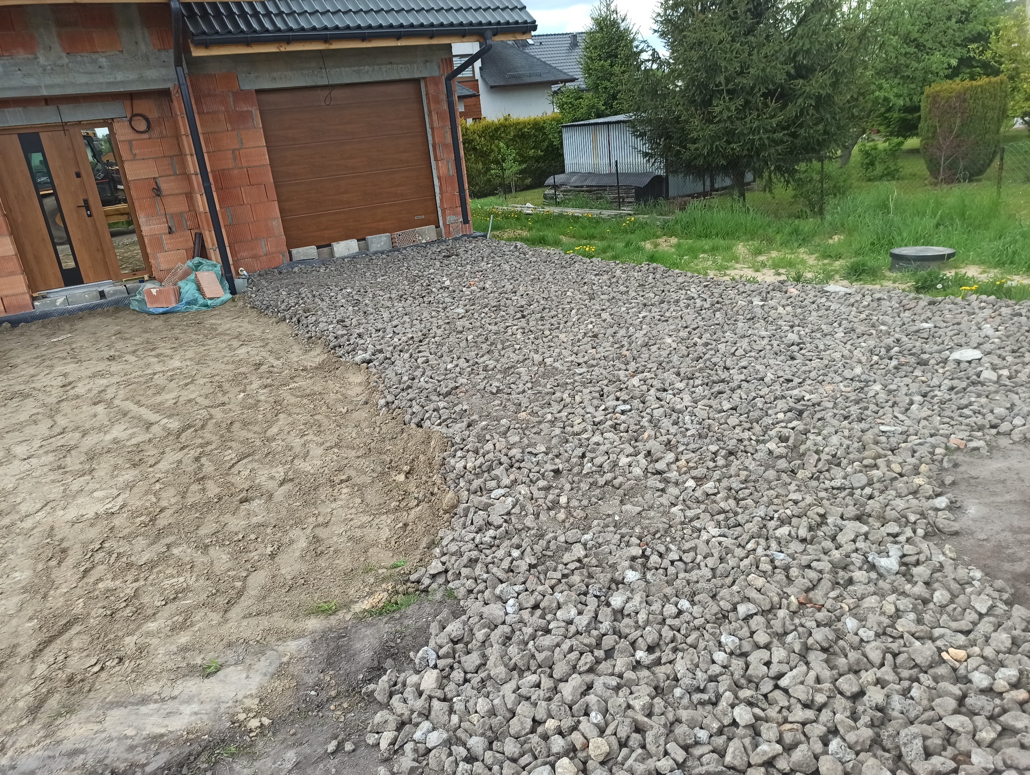 Przygotowanie terenu pod budowę: wysypany żwir na gruncie obok surowej bryły domu z cegły i brązową bramą garażową, widoczne nierówności terenu i pozostałości ziemi.