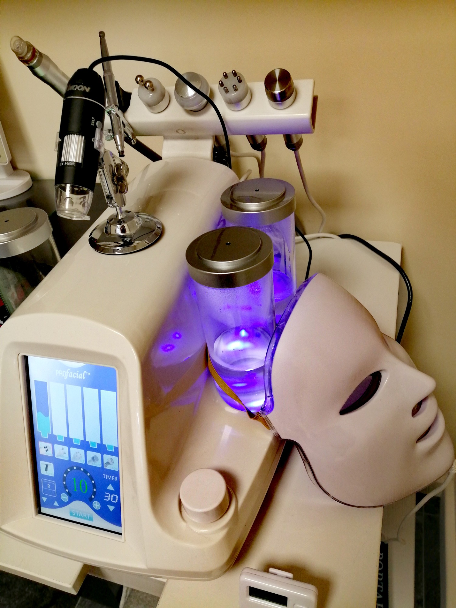Urządzenie do zabiegów kosmetycznych Profacial z maską LED, pojemnikami z płynem i mikroskopem cyfrowym, ustawione na beżowym blacie.