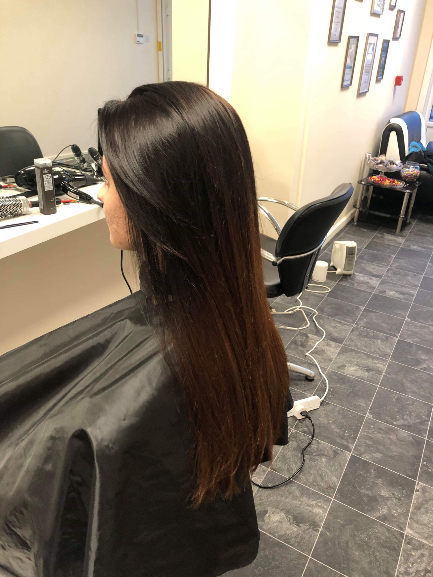 Kobieta siedzi w salonie z długimi, błyszczącymi, brązowymi włosami z ombre, ubrana w czarny fartuch fryzjerski, widoczny fragment blatu z akcesoriami i ścianą z dyplomami w tle.