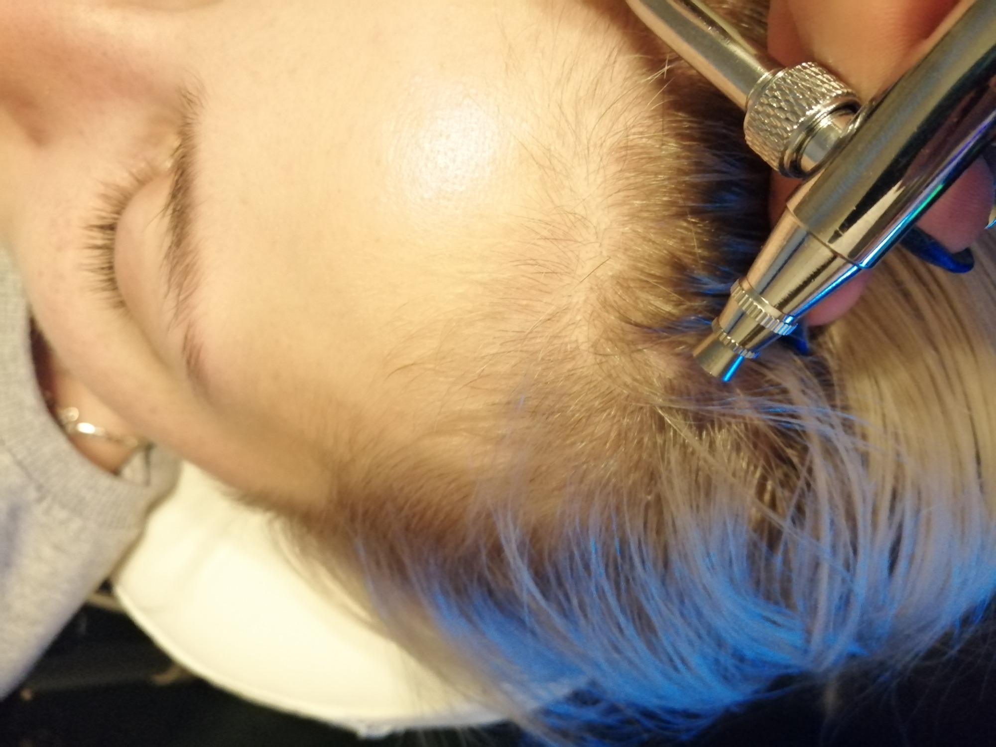 Aplikacja farby na odrosty włosów przy użyciu aerografu, widoczne dłonie z niebieskim manicure, włosy blond z odrostami.