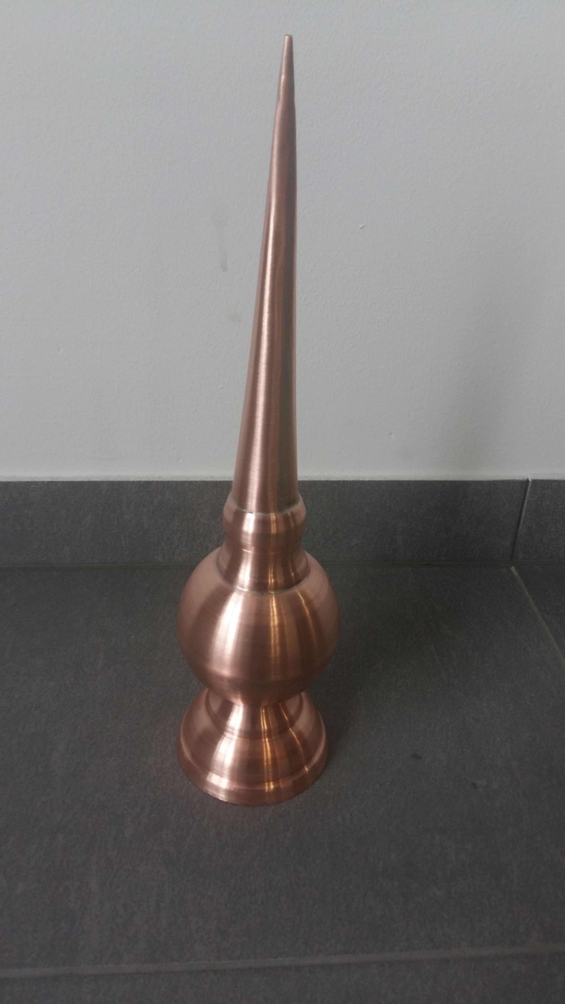 Miedziana iglica dachowa z dekoracyjną podstawą, stojąca na szarej podłodze na tle białej ściany.