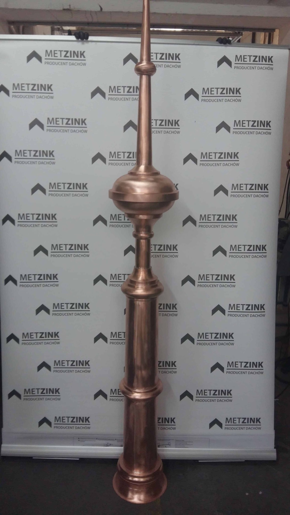 Miedziana iglica dachowa z dekoracyjnymi elementami, prezentowana na tle baneru reklamowego producenta pokryć dachowych.