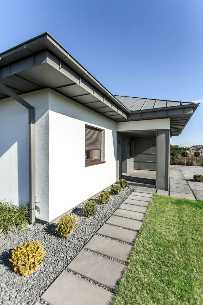 Nowoczesny dom jednorodzinny z szarą blachą na dachu i elewacją w kolorze złamanej bieli, widok z zewnątrz na wejście i ścieżkę z płyt betonowych.