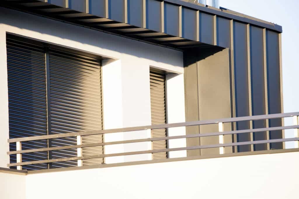 Nowoczesny budynek z ciemną blachą na dachu i elewacji, widok z balkonu z metalową balustradą, zamknięte żaluzje okienne.