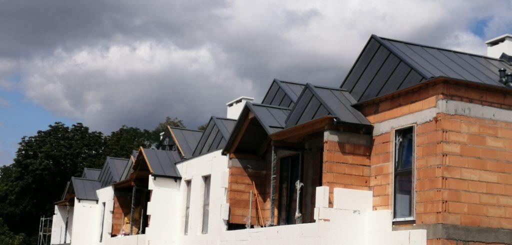 Szereg domów w trakcie budowy z nowymi, grafitowymi dachami z blachy na tle pochmurnego nieba.