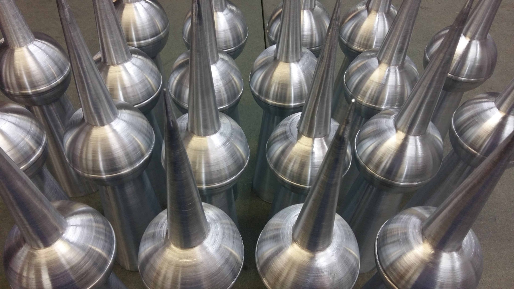 Szereg metalowych, polerowanych zakończeń kominowych w kształcie stożka z kulą, ułożonych na szarym tle, widoczne ślady obróbki mechanicznej.