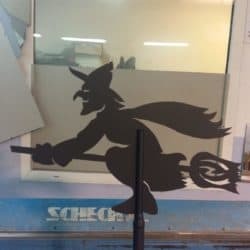 Sylwetka czarownicy na miotle, wycięta z blachy, umieszczona na tle okna z widocznym fragmentem napisu 'SCHECH'.