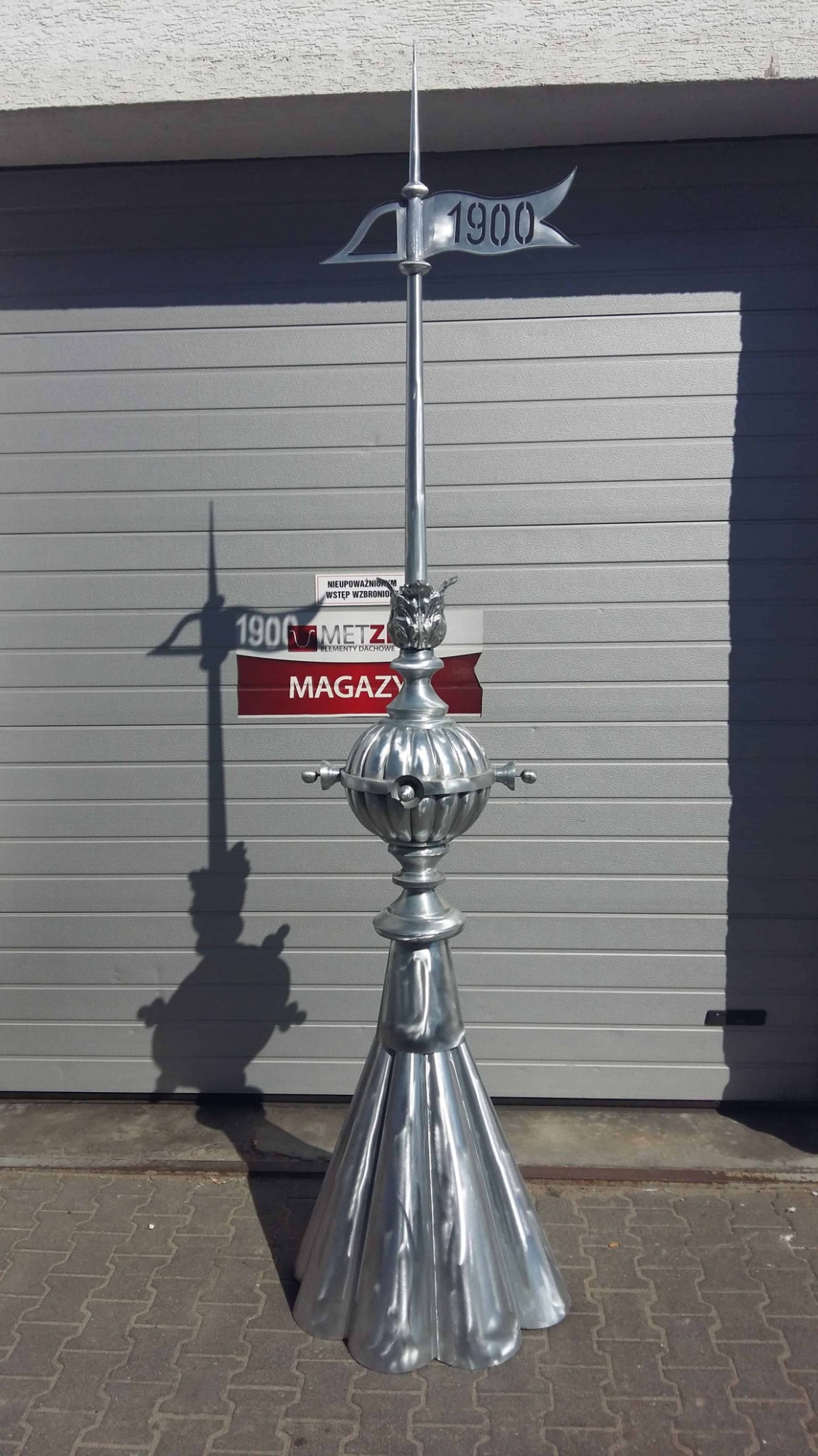 Metalowa iglica dachowa z datą 1900, umieszczona przed szarą bramą garażową, rzucająca cień.