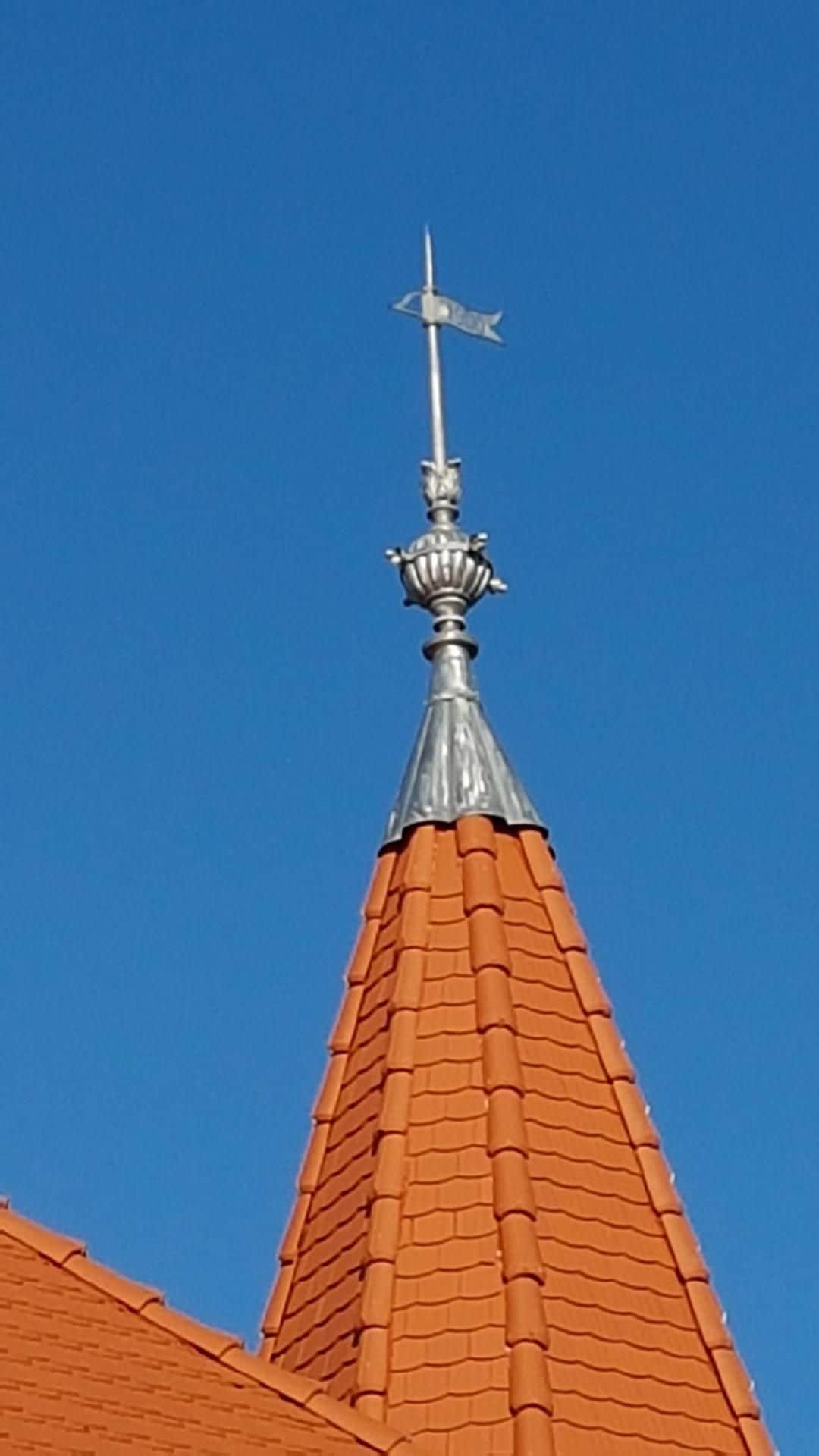 Wiatrowskaz z chorągiewką na szczycie wieży pokrytej czerwoną dachówką, zwieńczonej metalową obróbką blacharską, na tle błękitnego nieba.