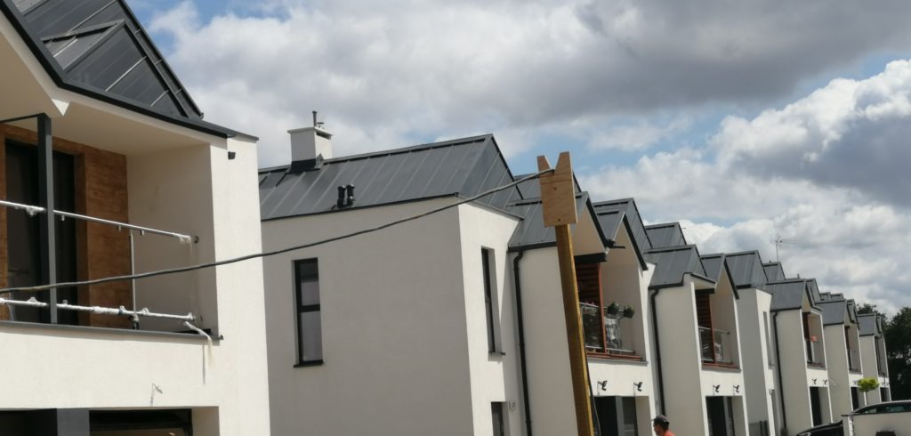 Szereg nowo wybudowanych, białych domów jednorodzinnych z ciemnymi dachami, widoczny z zewnątrz, z drewnianym słupem podtrzymującym przewody elektryczne przed jednym z domów.