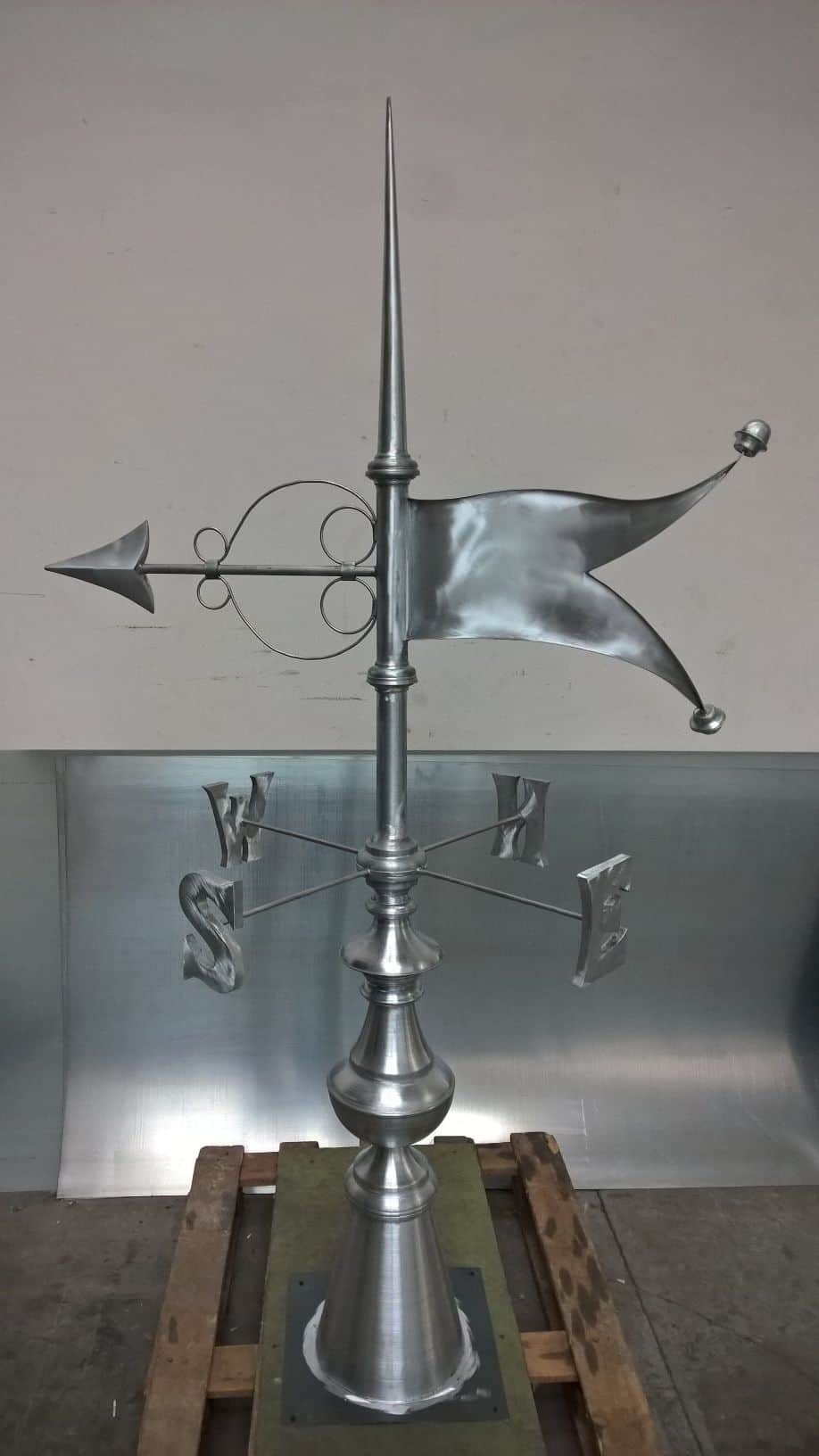 Metalowa chorągiewka wiatrowa z iglicą, strzałką, flagą i oznaczeniami kierunków świata, umieszczona na drewnianej palecie, w tle blaszane wykończenie ściany.