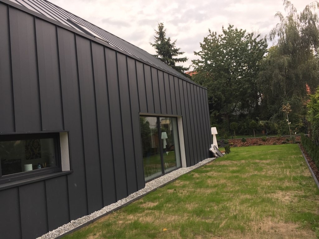 Nowoczesny dom z ciemną, metalową elewacją i obróbkami blacharskimi, widok z ogrodu, szara kolorystyka, widoczne okna i fragment dachu.