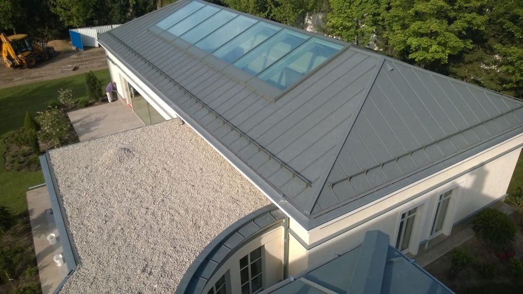 Szary dach z blachy z dużym świetlikiem na nowoczesnym domu, widok z góry, na posesji widoczna koparka.