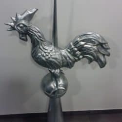 Metalowy kogut, wiatrowskaz na kuli, umieszczony na stożkowym wsporniku, z widocznymi detalami upierzenia i grzebienia.