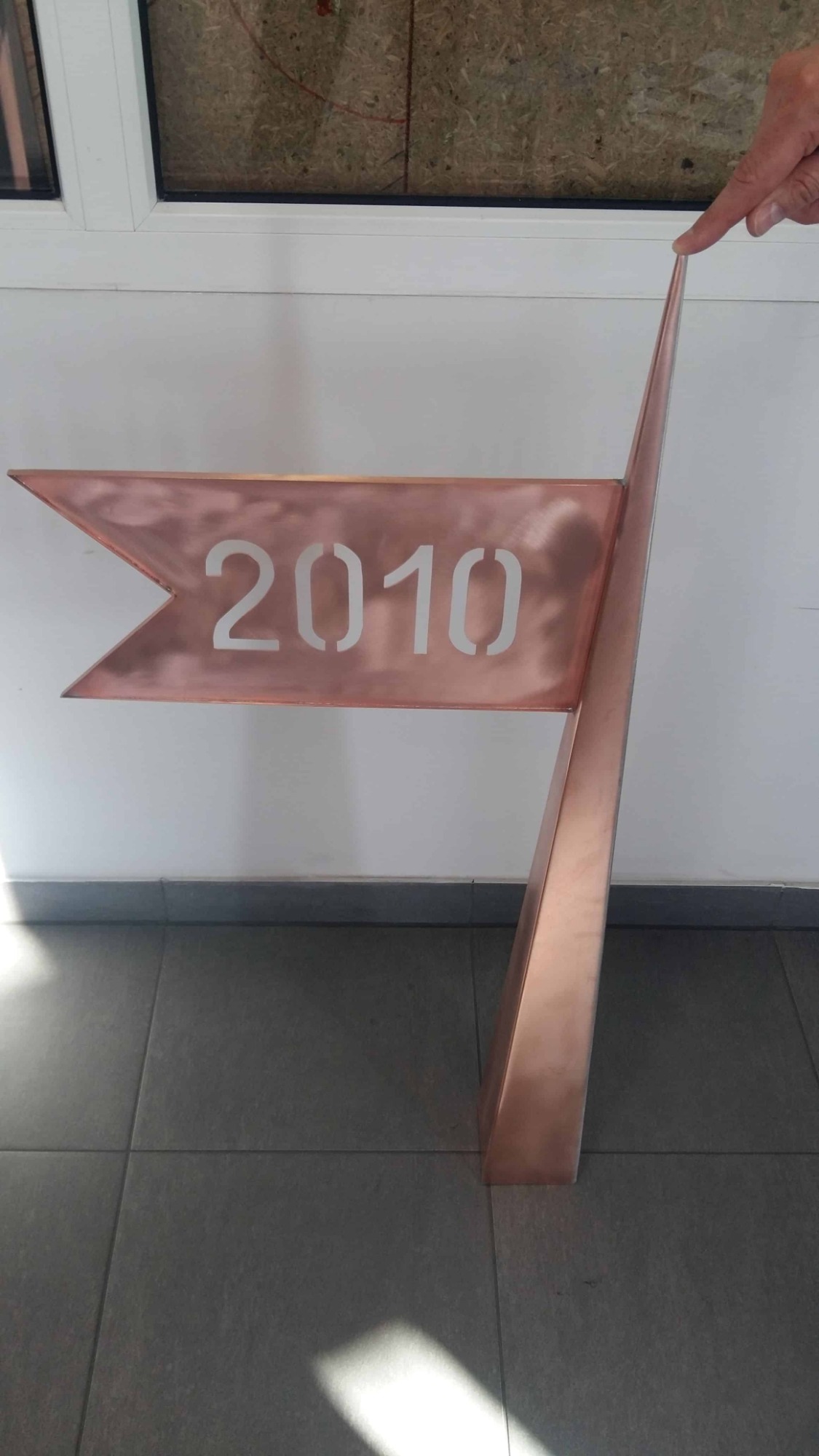 Miedziana flaga z wyciętym rokiem 2010, wskazana palcem, na tle białej ściany i szarej podłogi.