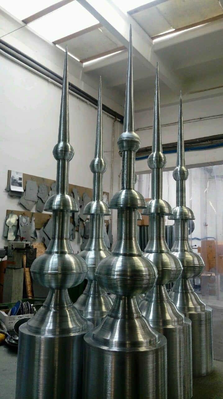 Szereg metalowych, obrobionych blacharsko elementów dekoracyjnych w kształcie wieżyczek z ostrymi zakończeniami, widoczne ślady obróbki mechanicznej, w tle warsztat blacharski.