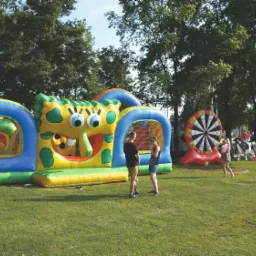 Rozległy dmuchany plac zabaw w kształcie SpongeBoba z tunelem z siatki, dmuchana tarcza do darta w tle, oraz osoby na trawiastym terenie.
