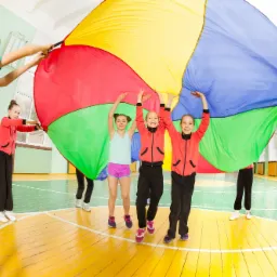 Grupa dzieci w sali gimnastycznej trzymająca wielokolorową chustę animacyjną, unosząc ją do góry podczas zabawy.