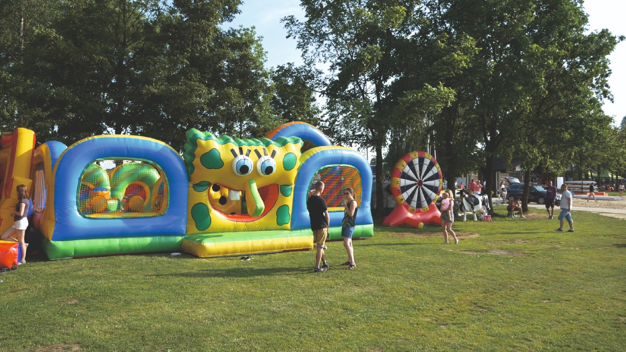 Rozległy dmuchany plac zabaw w kształcie SpongeBoba z tunelem z siatki, dmuchana tarcza do darta w tle, oraz osoby na trawiastym terenie.