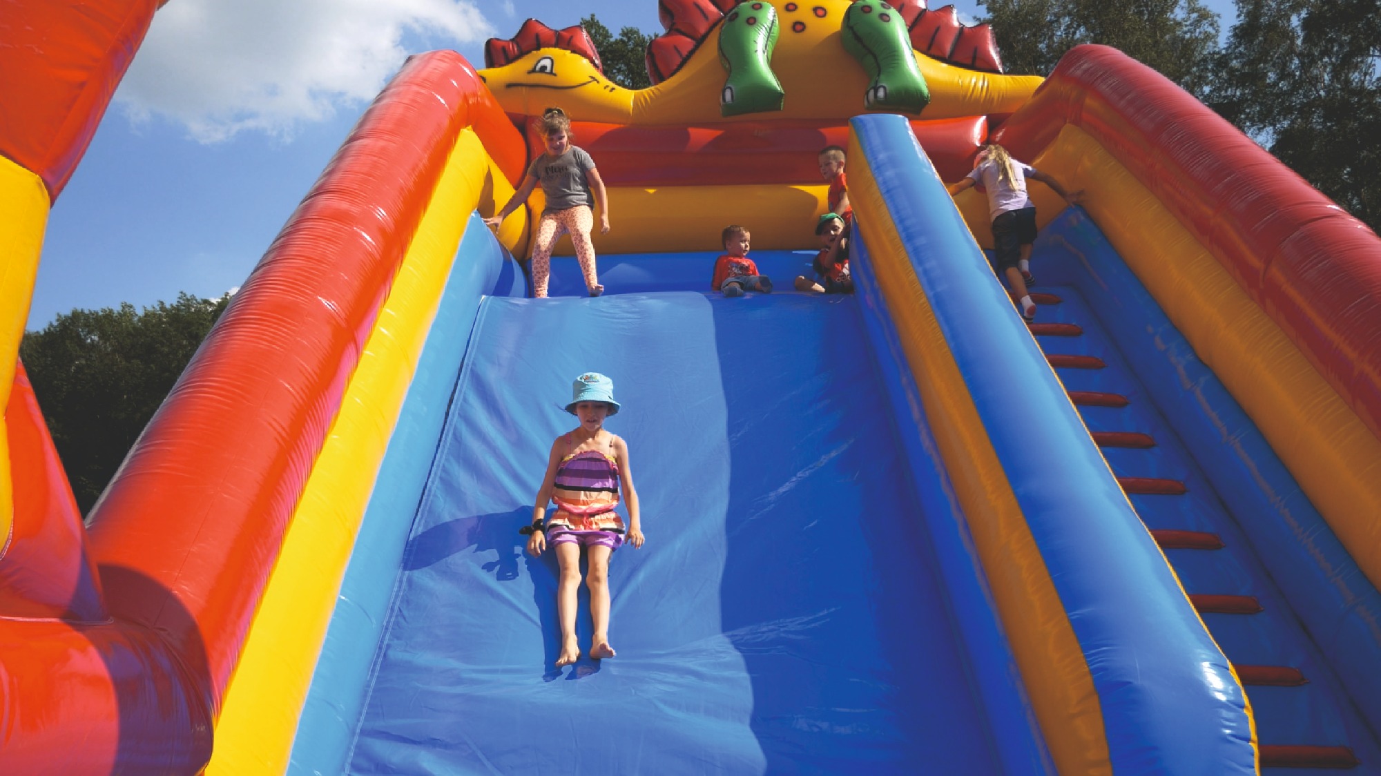 Dzieci na dmuchanej zjeżdżalni w kształcie dinozaura, dziewczynka w kapeluszu zjeżdża na dół, inne dzieci na górze i po bokach.