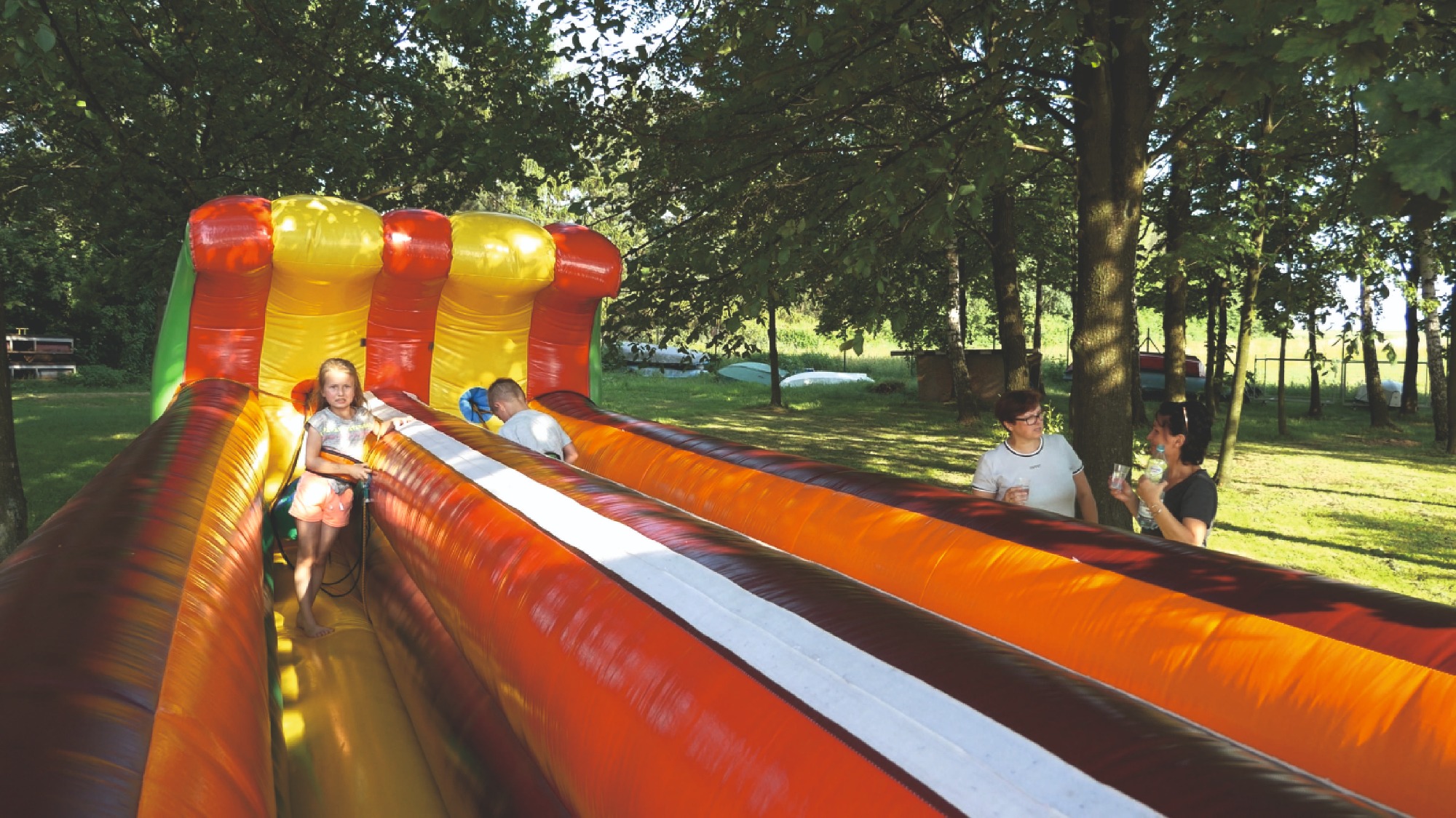 Dziewczynka wchodzi do dmuchanego tunelu z kolorowymi pasami na zielonym trawniku, w tle drzewa i dwie dorosłe osoby z napojami.