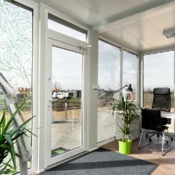 Wnętrze biura w module, z oknami ozdobionymi motywem liści, biurkiem, krzesłami i roślinami doniczkowymi, widok na plac budowy przez okno.