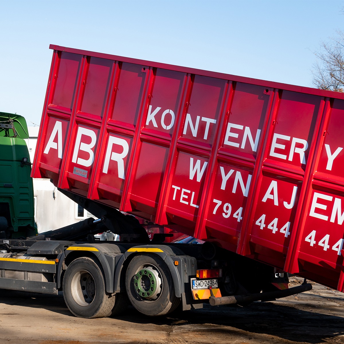 Czerwony kontener na gruz z białym napisem 'ABR KONTENERY WYNAJEM TEL 794 444 444' umieszczony na zielono-czarnej ciężarówce z podniesioną skrzynią, widok z boku w słoneczny dzień.