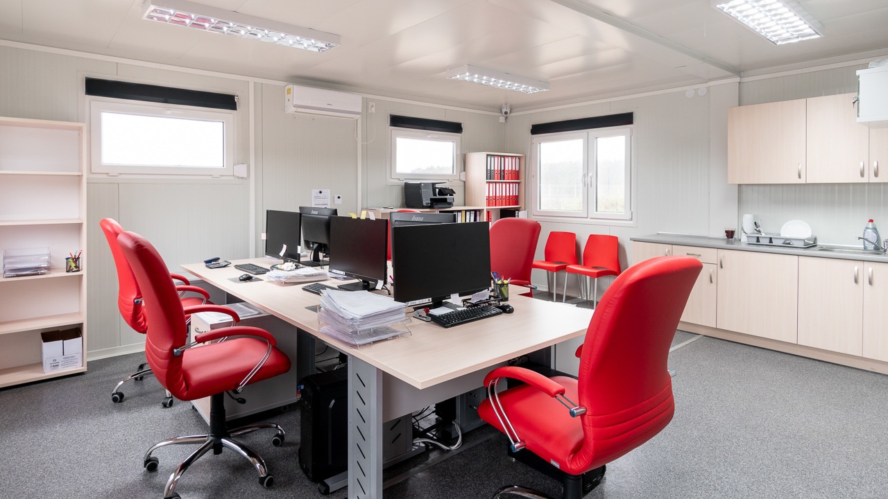 Wnętrze biura w domu modułowym z jasnymi ścianami, szarym dywanem, biurkami z komputerami i czerwonymi krzesłami obrotowymi oraz aneksem kuchennym.