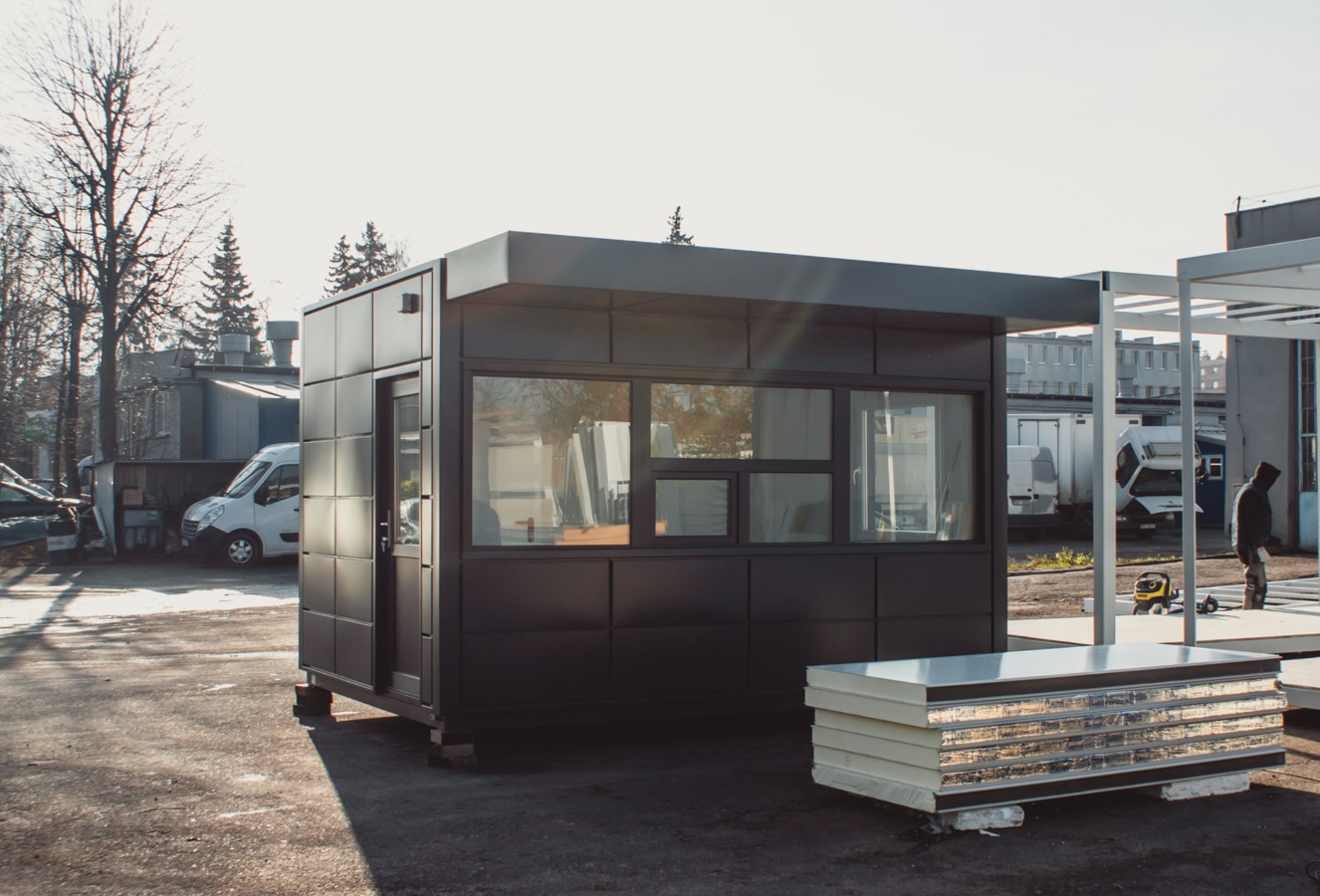 Czarny, nowoczesny pawilon modułowy z dużymi oknami, widok z zewnątrz na placu budowy, obok leżą panele izolacyjne.