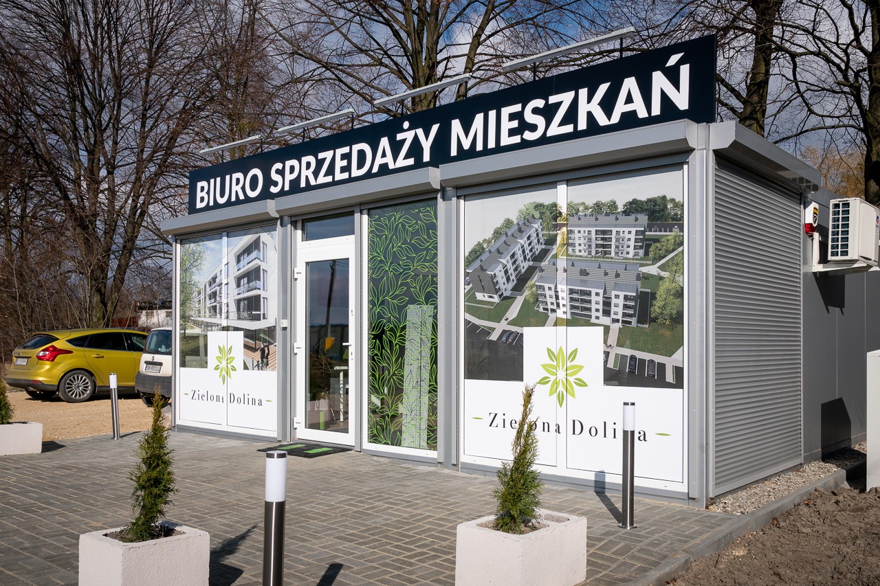 Biuro sprzedaży mieszkań 'Zielona Dolina' w Mikołowie, z wizualizacjami budynków na oknach i szyldem 'Biuro Sprzedaży Mieszkań' nad wejściem, przed budynkiem zaparkowane dwa samochody.