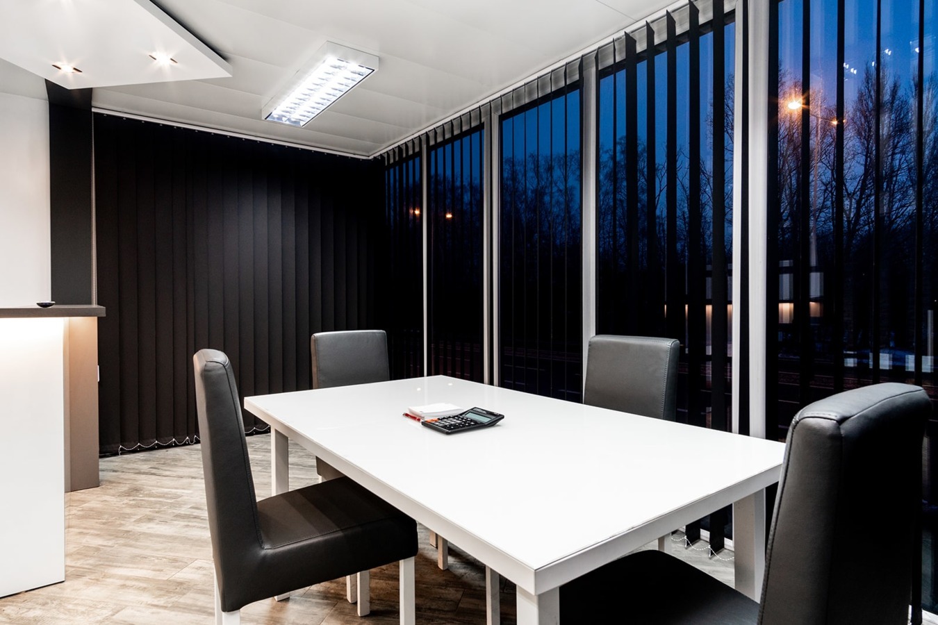 Wnętrze biurowe w module: biały stół, czarne krzesła, kalkulator i notatnik na blacie, okna z pionowymi żaluzjami i widok na nocny krajobraz.