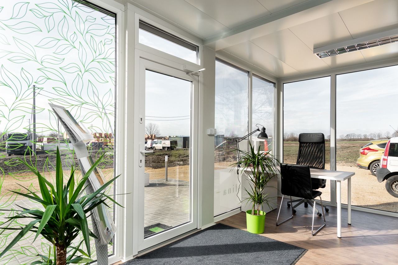 Wnętrze biura w module, z oknami ozdobionymi motywem liści, biurkiem, krzesłami i roślinami doniczkowymi, widok na plac budowy przez okno.