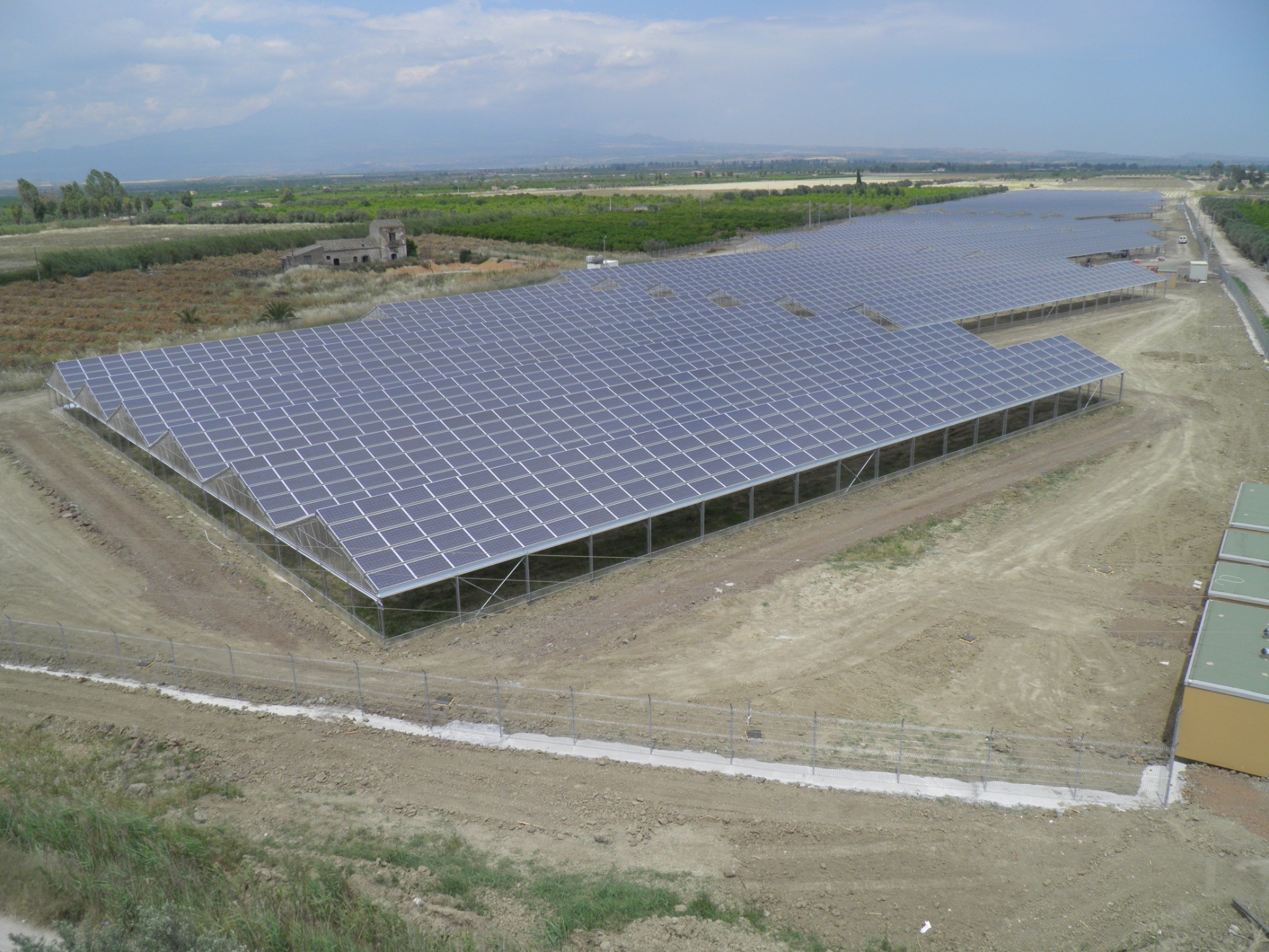 Rozległa farma paneli słonecznych na konstrukcji stalowej, otoczona ogrodzeniem z drutu kolczastego, z widokiem na górzysty krajobraz w tle.