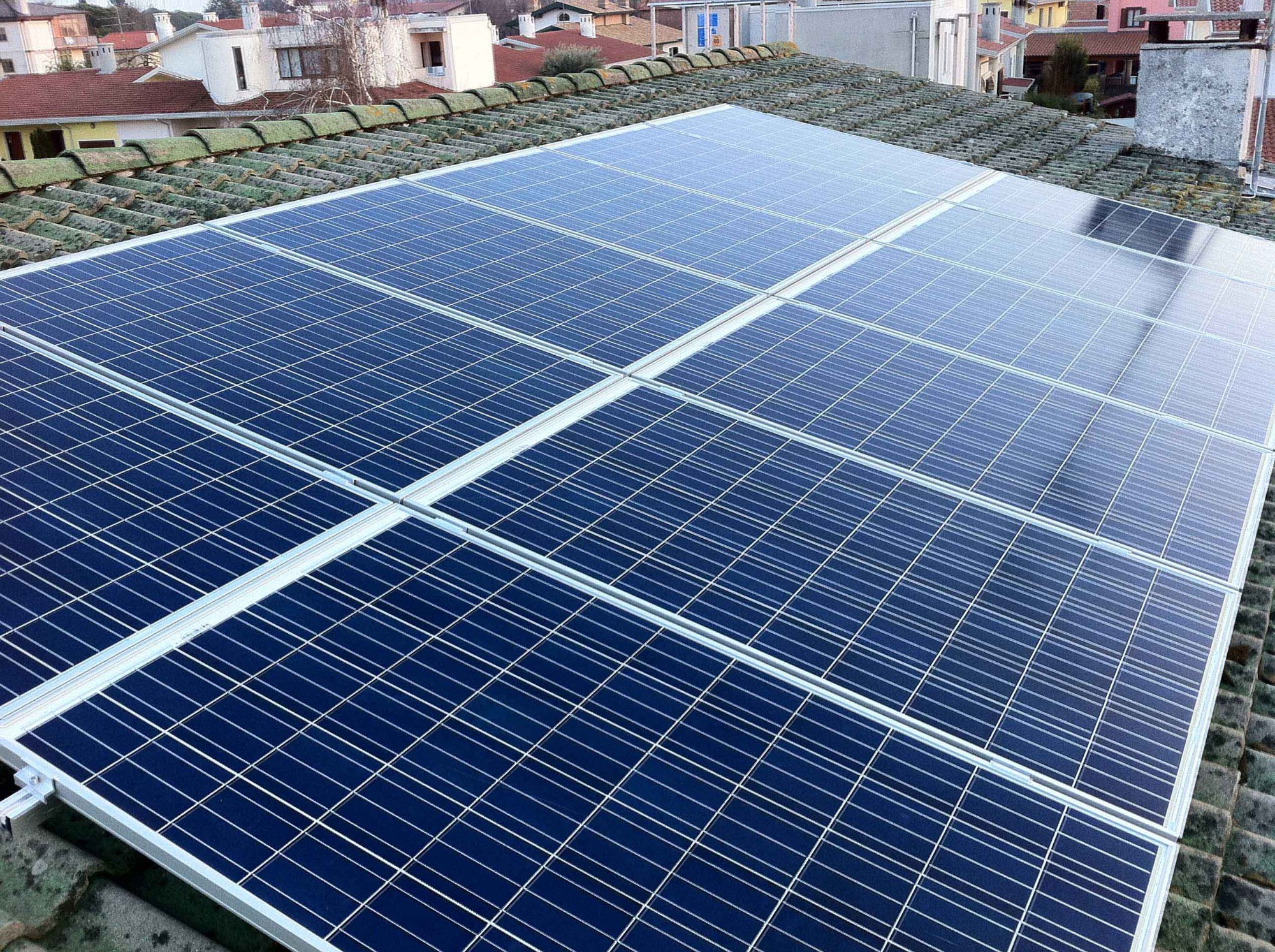 Panele słoneczne na dachu z dachówką pokrytą mchem, widoczne budynki w tle.